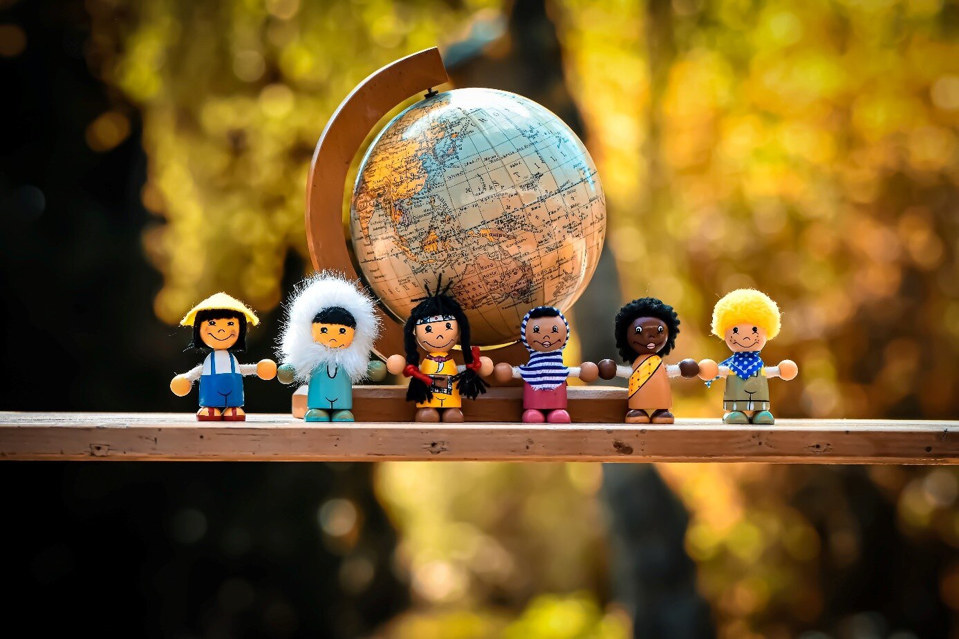 Auf einem Brett steht im Hintergrund ein Globus. Davor stehen sechs Puppen mit unterschiedlicher Hautfarbe und unterschiedlicher Kleidung. Sie halten sich an den Händen.